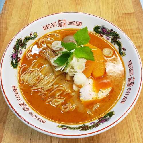 A bowl of rich, orange curry noodle soup topped with vegetables, herbs, and a soft-boiled egg on a wooden surface.