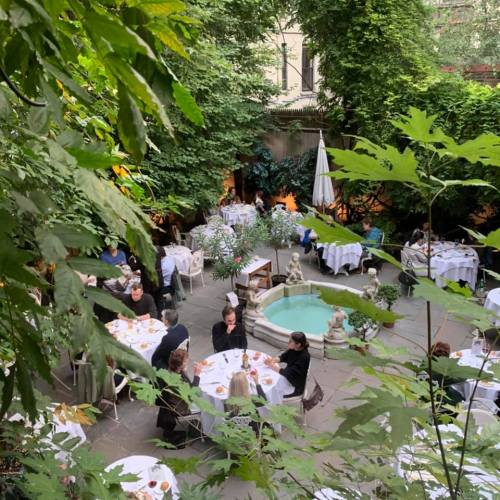A lush outdoor dining area with tables set for guests, surrounded by greenery and a small fountain.