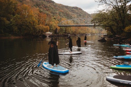 Four people in black cloaks paddle on stand-up boards in a river surrounded by autumn foliage and a bridge in the background.