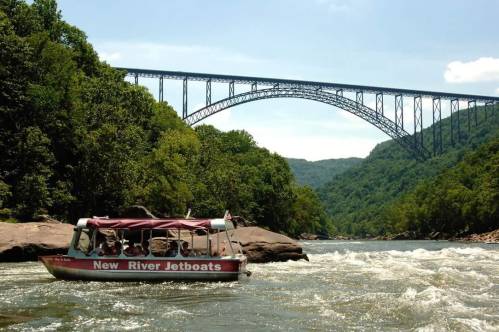 A jetboat navigates the river beneath a large steel arch bridge, surrounded by lush green hills and a clear blue sky.