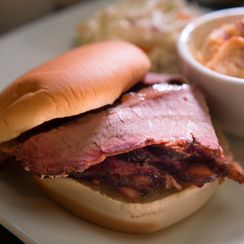 A close-up of a sandwich with thick slices of smoked brisket on a soft bun, served with coleslaw and a side dish.