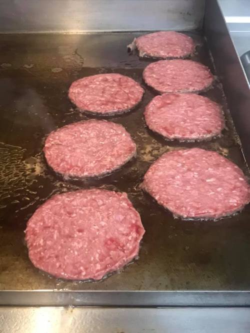 Seven raw hamburger patties sizzling on a griddle, surrounded by oil.