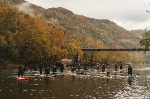 A group of people in wetsuits paddleboarding on a river surrounded by autumn foliage and a bridge in the background.