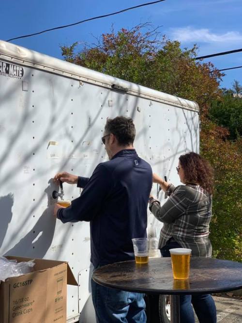 Two people attach items to a white trailer while drinks sit on a table nearby, surrounded by greenery.