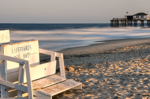 A lifeguard chair on a sandy beach with a pier in the background and gentle waves under a clear sky.