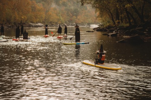 A group of people in witch costumes paddleboarding on a serene river surrounded by autumn foliage.