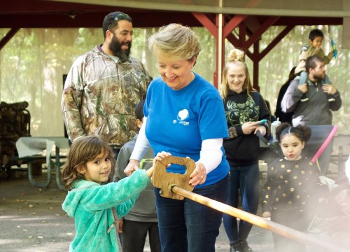 A woman in a blue shirt helps a young girl with a wooden toy, surrounded by smiling adults and children in a park setting.