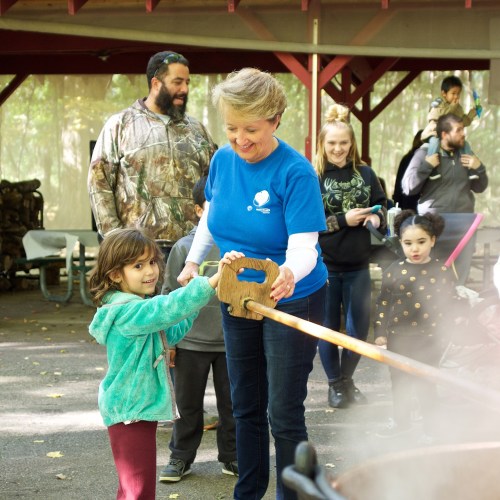 A woman and a young girl stir a large pot outdoors, surrounded by smiling people in a forested setting.
