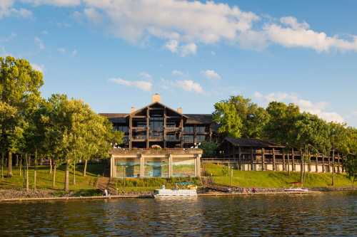 A lakeside lodge surrounded by trees, featuring a large deck and a boat dock under a clear blue sky.