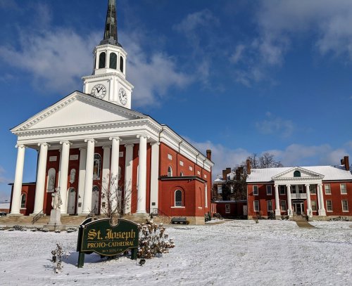 St. Joseph Proto-Cathedral, a red brick building with a clock tower, surrounded by snow and a clear blue sky.