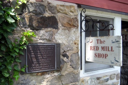 A stone wall with a plaque and a sign reading "The Red Mill Shop," surrounded by greenery.