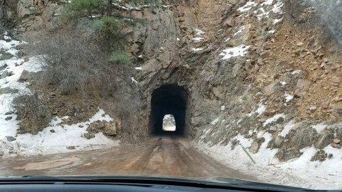 A dirt road leads through a rocky tunnel, with snow and vegetation on either side. Light shines at the tunnel's end.