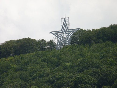 A large metal star structure atop a green hillside, surrounded by trees under a cloudy sky.