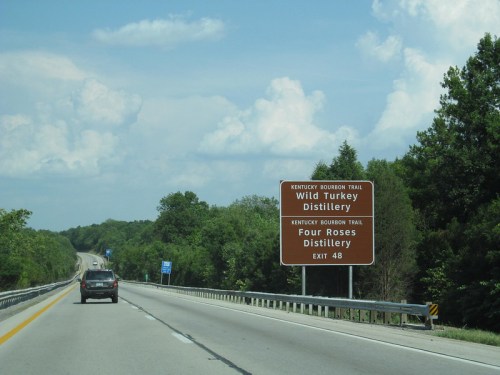 Road sign for Wild Turkey and Four Roses distilleries on the Kentucky Bourbon Trail, with a highway and trees in the background.