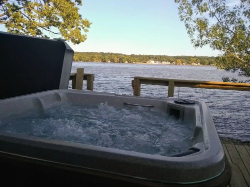 A hot tub on a deck overlooking a calm lake, surrounded by trees and a clear blue sky.