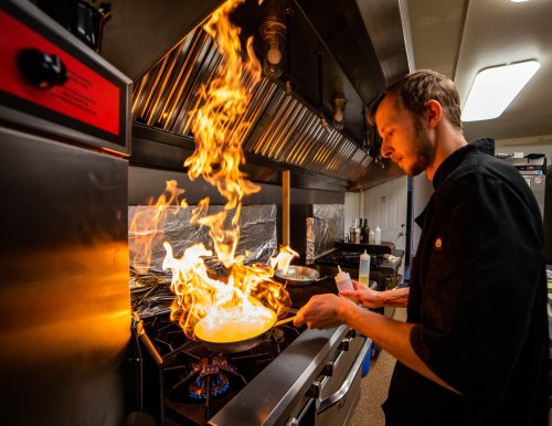 A chef cooks in a kitchen, flames rising dramatically from a pan on the stove.