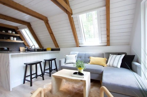 Cozy living room with sloped wooden beams, a gray sofa, a small coffee table, and a kitchenette in a bright, airy space.