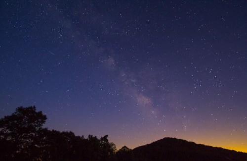 A starry night sky with the Milky Way visible above a silhouette of a mountain and trees.