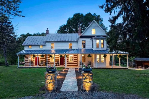 A charming two-story house with a metal roof, surrounded by greenery and illuminated pathways at dusk.