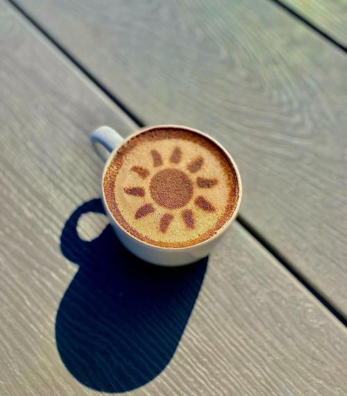 A cup of coffee with a sun design on the frothy surface, resting on a wooden table.