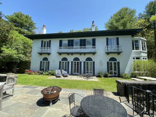 A charming two-story house with blue shutters, balconies, and a patio surrounded by greenery and a fire pit.