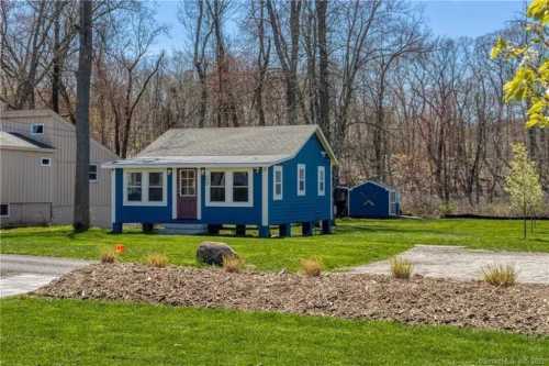 A small blue house with a porch surrounded by green grass and trees, with a gravel path and a shed in the background.