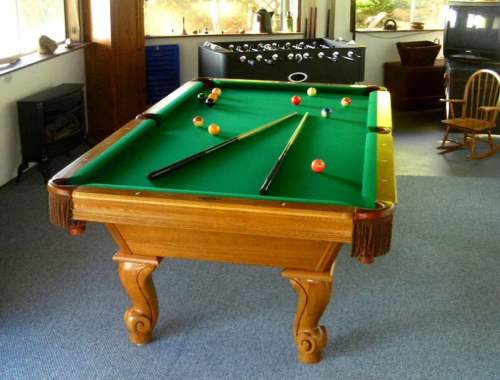 A wooden pool table with green felt and balls, set in a cozy room with a foosball table and a rocking chair.