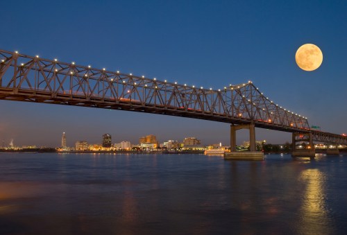 A bridge illuminated at night with a large moon overhead and a city skyline reflecting in the water below.