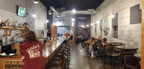 A cozy restaurant interior with a long bar, patrons dining, and a TV on the wall. Warm lighting and rustic decor.