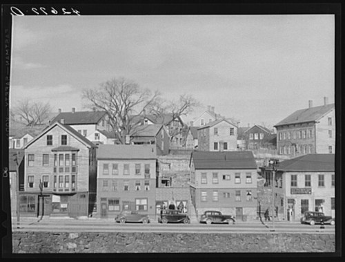 Historic black-and-white photo of a row of houses and cars along a street, with trees and hills in the background.