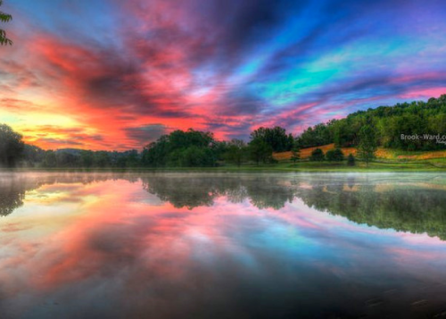 Vibrant sunset over a calm lake, with colorful clouds reflecting on the water and lush greenery in the background.