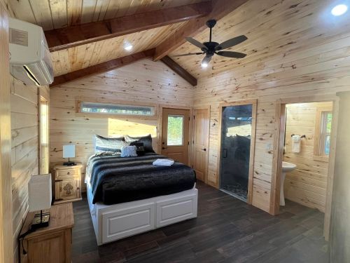 Cozy wooden bedroom with a king-sized bed, ceiling fan, and natural light from windows. Modern rustic decor.