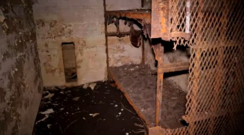 A dimly lit, abandoned room with peeling walls and a rusty metal shelf, suggesting neglect and decay.