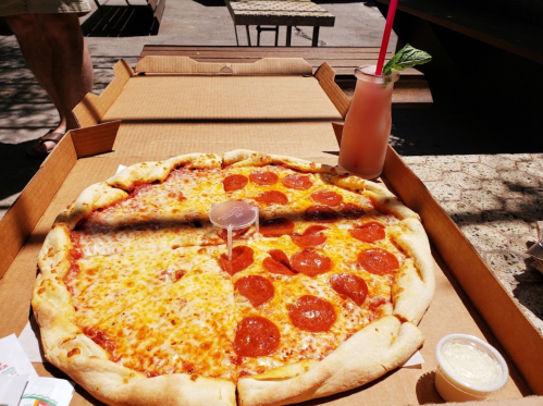 A large pizza with half cheese and half pepperoni, alongside a pink drink with a mint leaf, on a picnic table.