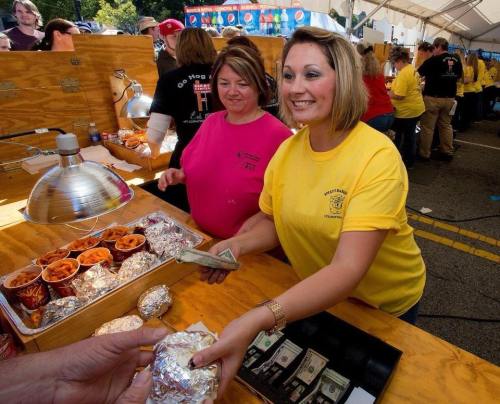 Two women serve food at a festival booth, smiling as one hands over a wrapped item while the other collects payment.