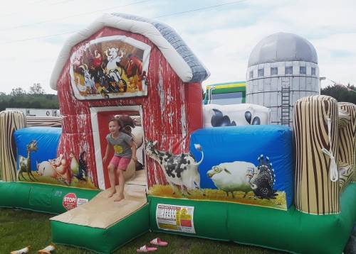 A child joyfully exits a colorful inflatable barn with animal graphics, set in a grassy area under a blue sky.