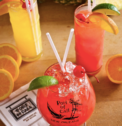 Three colorful cocktails with fruit garnishes, including a vibrant red drink in a branded glass, on a wooden table.