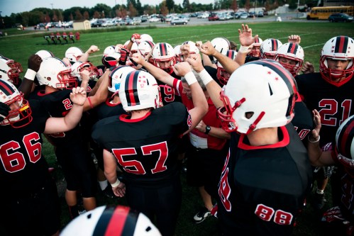 A group of football players in black jerseys huddle together, raising their hands in a show of team spirit.