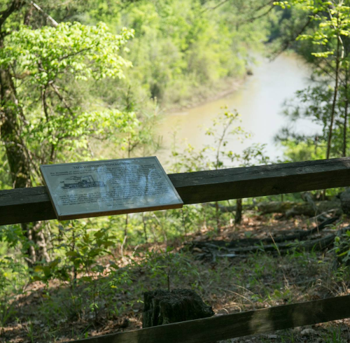 A sign on a wooden fence overlooking a river surrounded by lush greenery and trees.