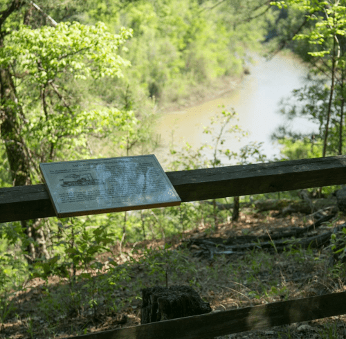 A sign on a wooden fence overlooking a river surrounded by lush greenery and trees.