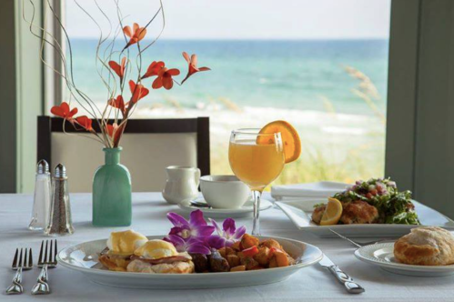 A beautifully set table with breakfast, a mimosa, and flowers, overlooking a beach view.
