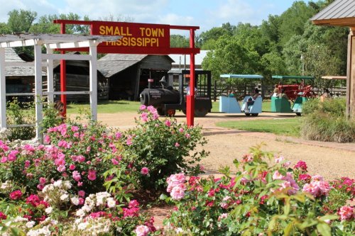 A vibrant garden with roses leads to a sign reading "Small Town Mississippi," with vintage trains in the background.