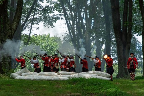 Historical reenactors in red uniforms fire muskets amidst smoke, surrounded by trees and sandbags.