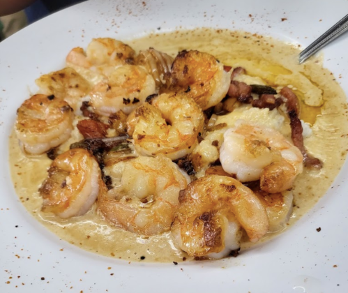 A plate of shrimp cooked in a creamy sauce, garnished with herbs and spices, served with a side of crispy bacon.