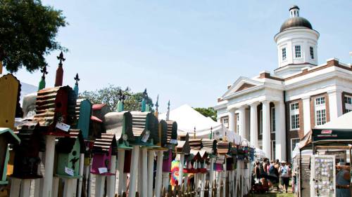 Colorful birdhouses line a pathway at an outdoor event, with a historic building and tents in the background.