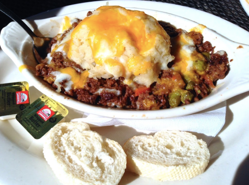 A dish of shepherd's pie topped with melted cheese, served with butter packets and slices of bread on the side.