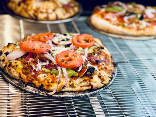 A freshly baked pizza topped with tomatoes, onions, and green peppers on a wire rack, with more pizzas in the background.