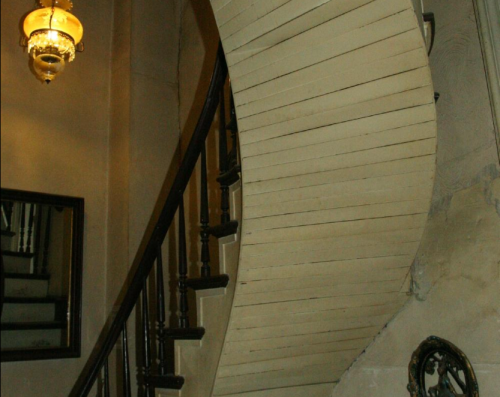 A curved wooden staircase with a vintage chandelier and a mirror reflecting the steps.
