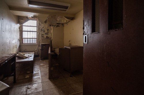 A dimly lit, abandoned room with peeling walls, a barred window, and scattered debris on the floor.