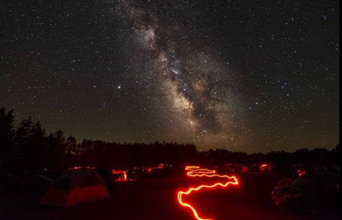 A starry night sky with the Milky Way above a campsite, illuminated by red lights from tents and pathways.